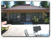 Outdoor patio area at Park Folsom Senior Retirement Community featuring a paved concrete surface, a bench on the left, a round table with chairs in the foreground, and a building with large windows and a tiled roof in the background surrounded by greenery.