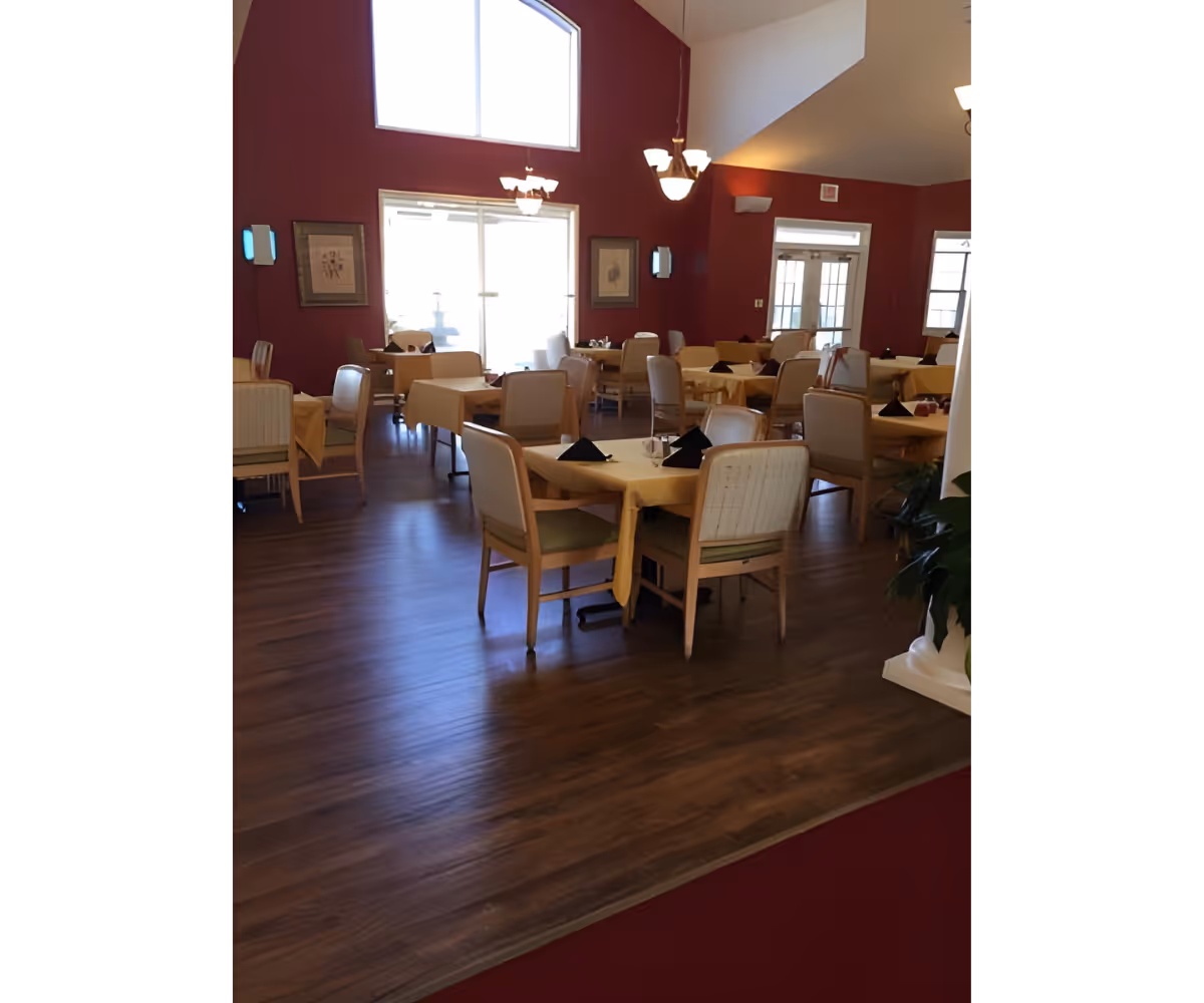 Interior view of a dining room with multiple tables covered with yellow tablecloths and set with black napkins and glassware. The room has wooden flooring, red walls, large windows, and ceiling lights hanging from a high ceiling.