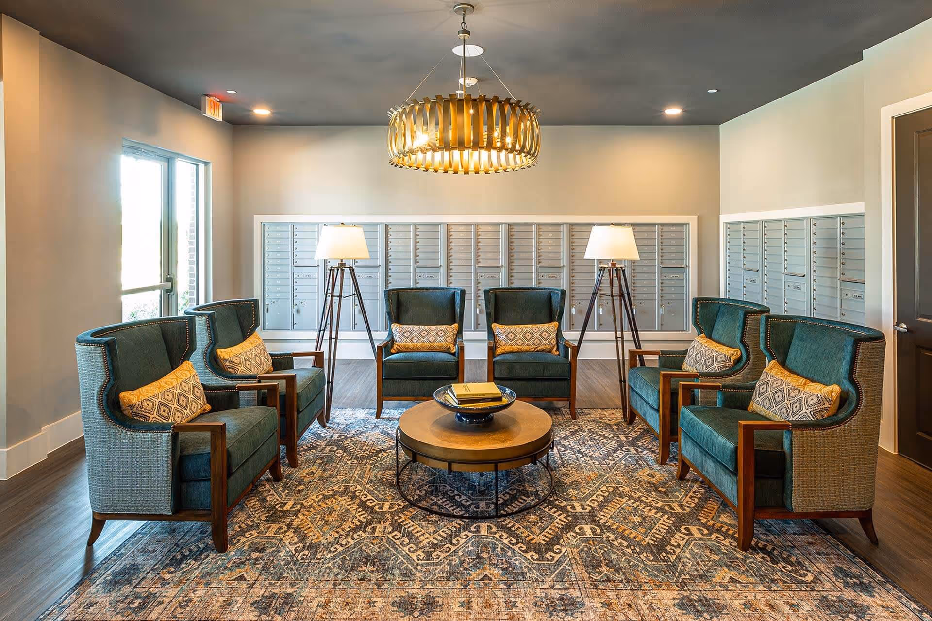 A cozy seating area with six green upholstered armchairs arranged around a round wooden coffee table on a patterned rug. Two floor lamps with white shades stand behind the chairs, and a modern circular chandelier hangs from the ceiling. The back wall features multiple mailboxes, and there is a glass door to the left letting in natural light.