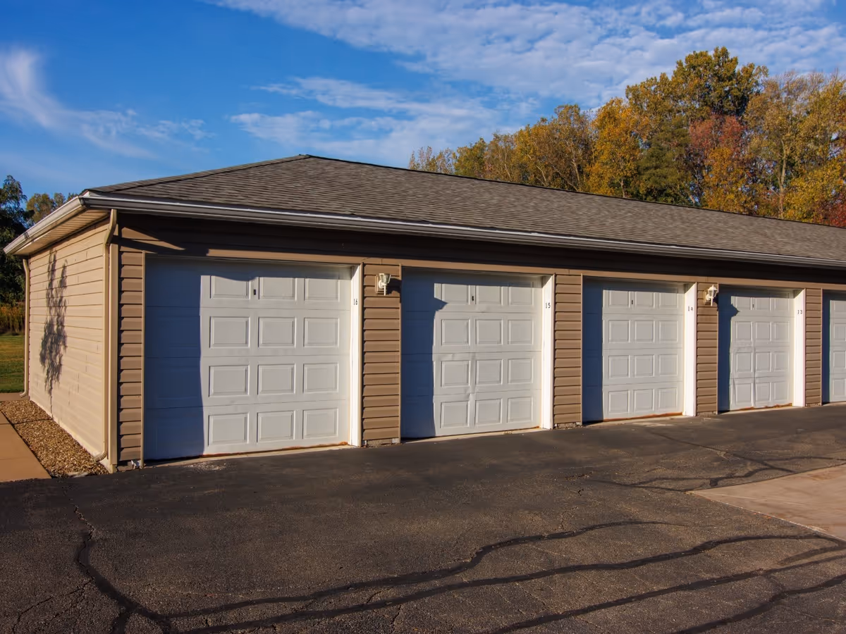 Row of beige garage units with white doors along a paved drive and autumn trees.