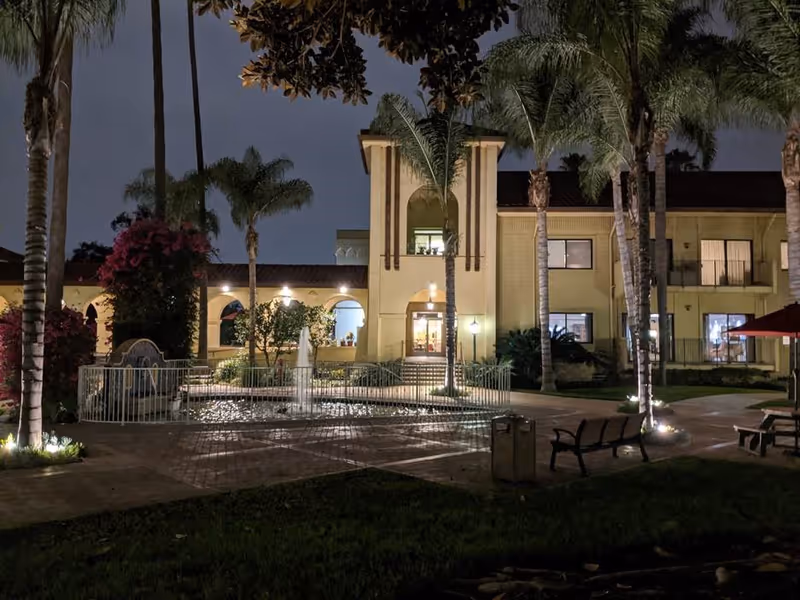 Night view of Hollenbeck Palms courtyard showing a lit fountain, palm trees, benches, and the illuminated building entrance.