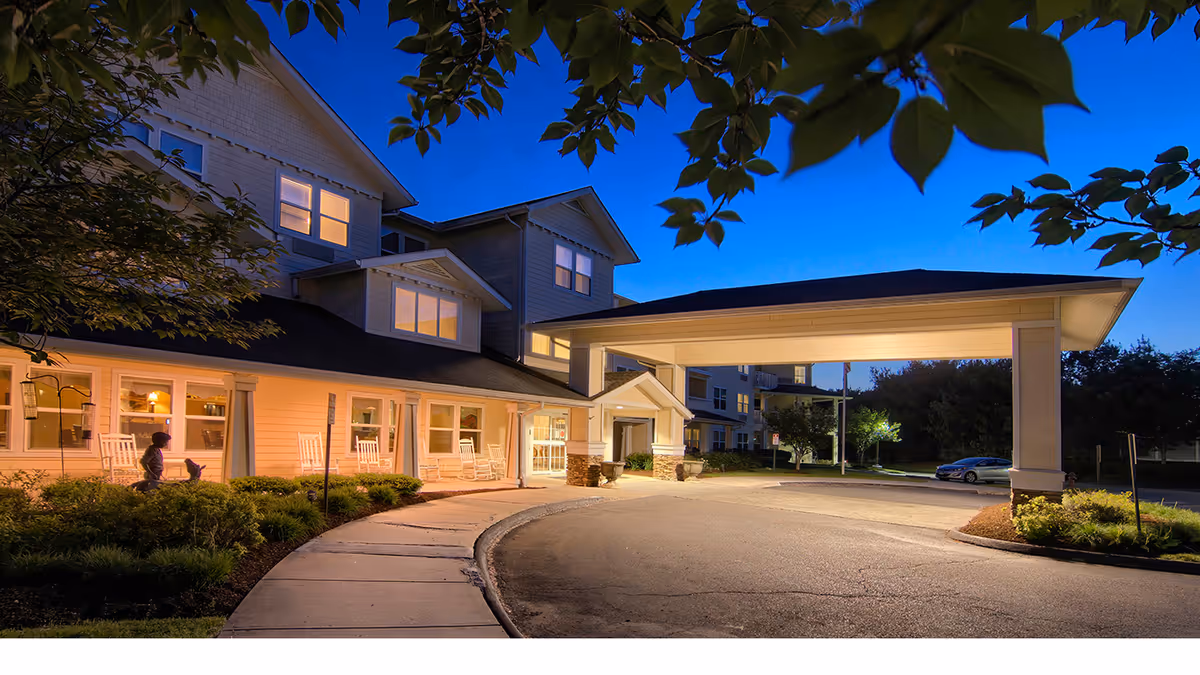 Front entrance of a senior living building at dusk with a covered porte-cochère, lit windows, and rocking chairs on the porch.