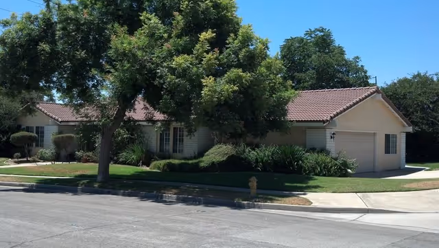 Single-story beige senior living facility with red-tiled roof, manicured lawn, and driveway along street.