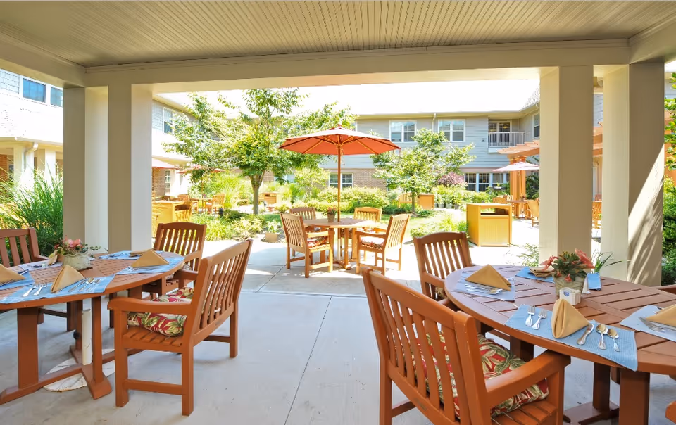 Outdoor patio area at The Cliffs At Eagle Rock with wooden tables and chairs, some tables set with placemats, napkins, and utensils. There are umbrellas providing shade and greenery including trees and bushes surrounding the seating area.