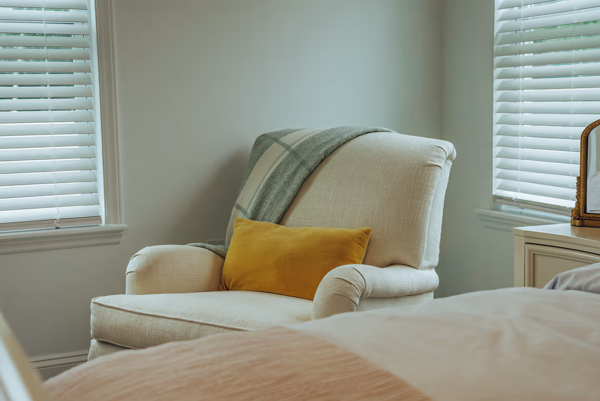 A cream armchair with a yellow pillow sits beside a bed in a bright bedroom.