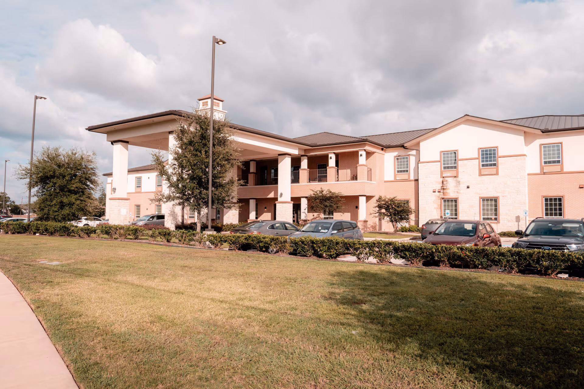 Front exterior of a two-story senior living facility with a covered entrance, parked cars, and landscaped lawn.