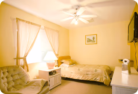 Cozy bedroom in a senior facility with bed, nightstand, lamp, and chair.