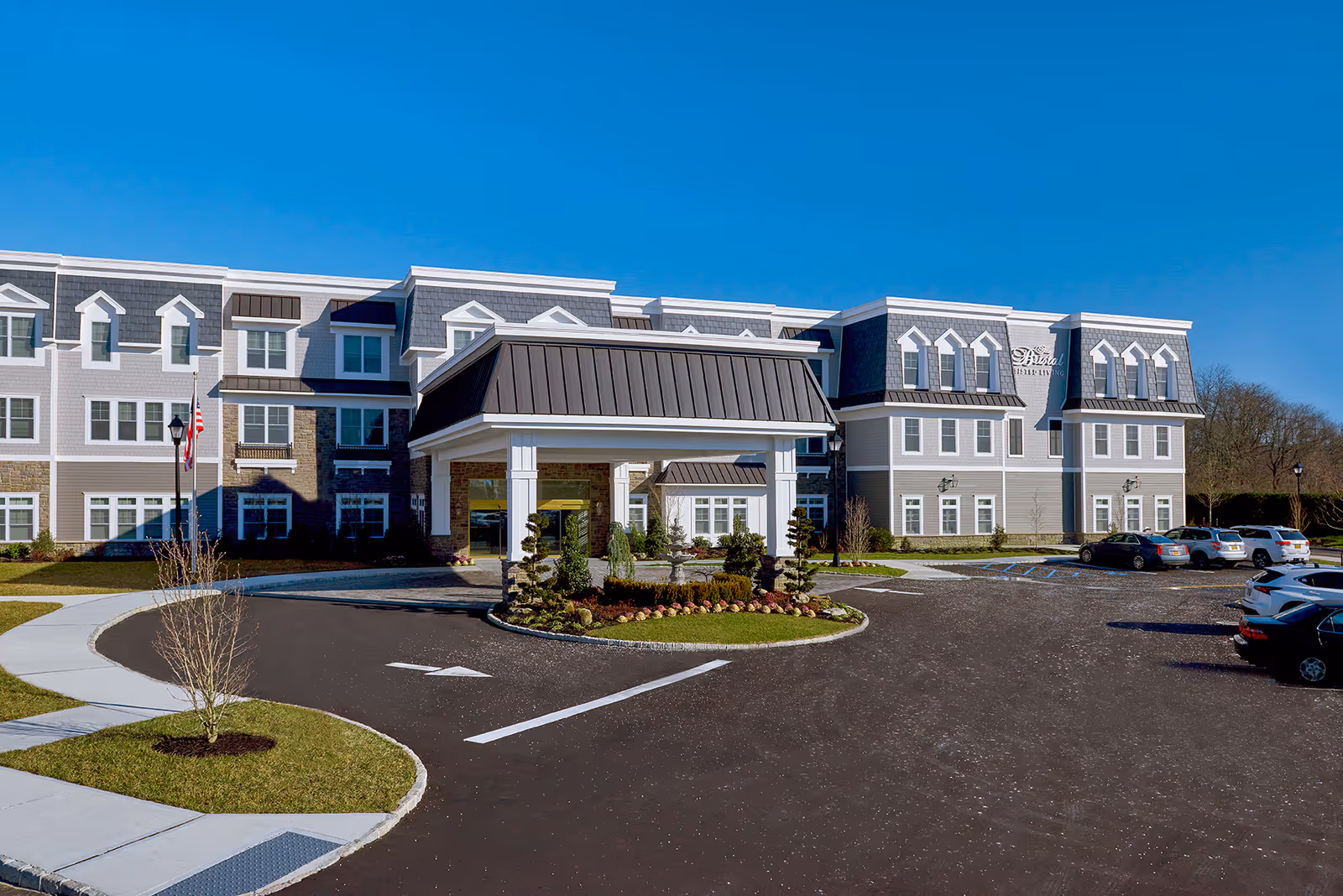 Three-story assisted living building with a covered porte-cochère, circular driveway, landscaping, and parked cars under a clear blue sky.