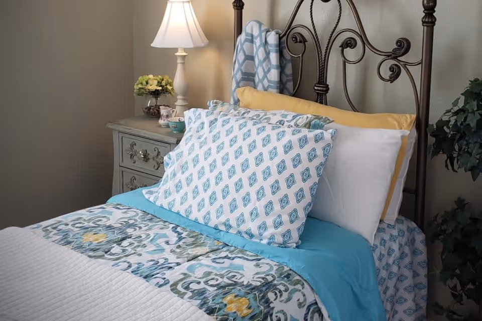 A neatly made bed with patterned blue and white pillows and a matching quilt. The bed has a decorative metal headboard. Next to the bed is a vintage-style nightstand with a white lamp, a small flower arrangement, and a teacup. A green leafy plant is visible on the right side of the image.