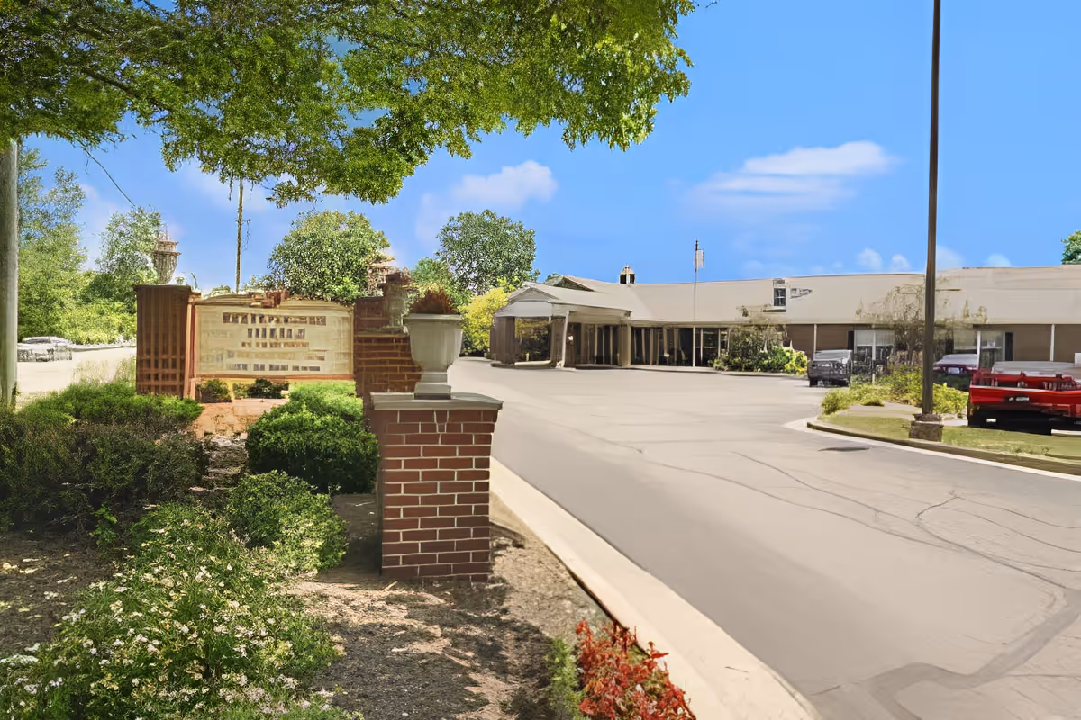 Front entrance and driveway of a single-story senior living facility with a brick sign, landscaped beds, and parked cars.