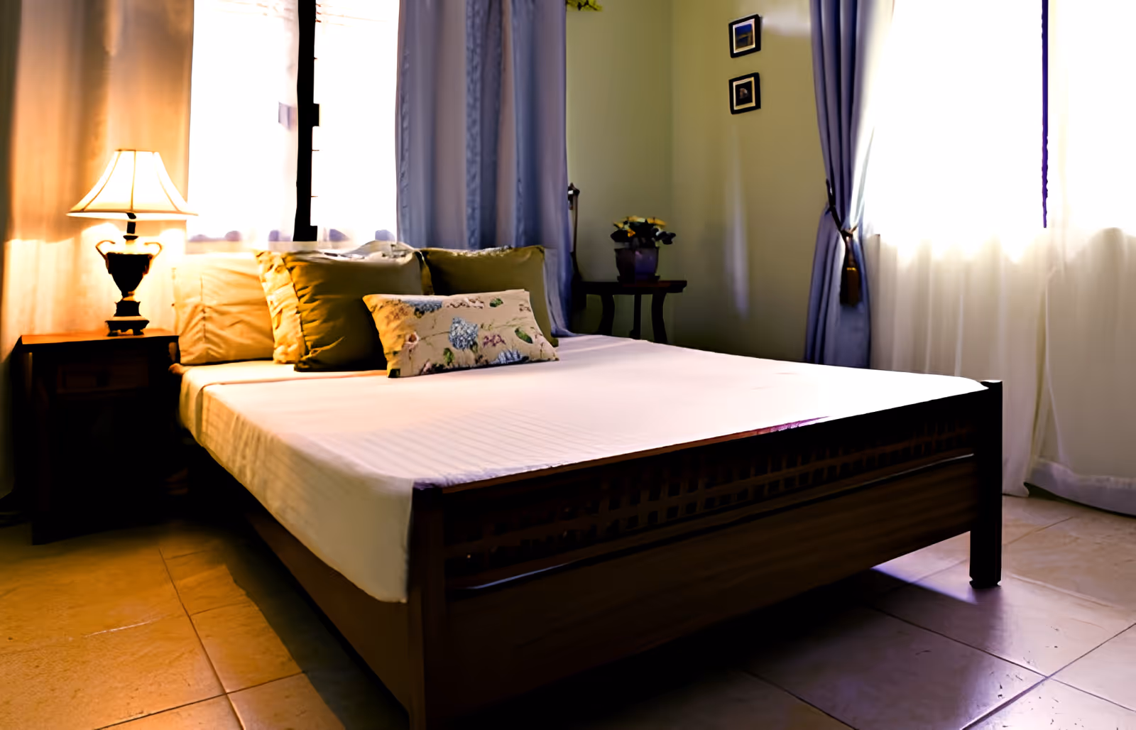 A neatly made bed with multiple pillows in a softly lit bedroom. There is a wooden nightstand with a lamp on the left side and a small table with a potted plant in the corner. Light blue curtains frame two windows, allowing natural light to brighten the room.