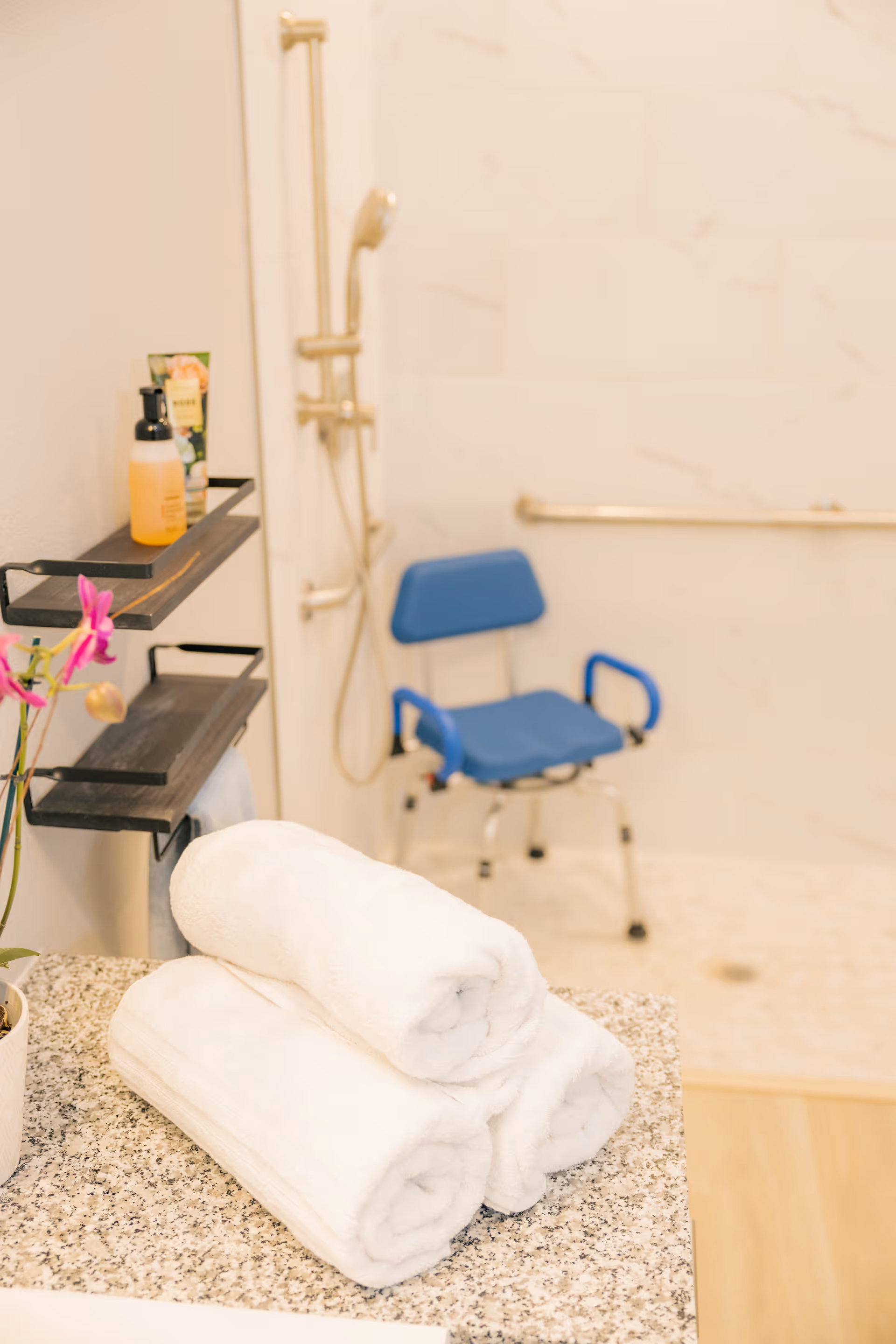 Bright bathroom with blue shower chair, grab bars, and folded towels on granite counter.