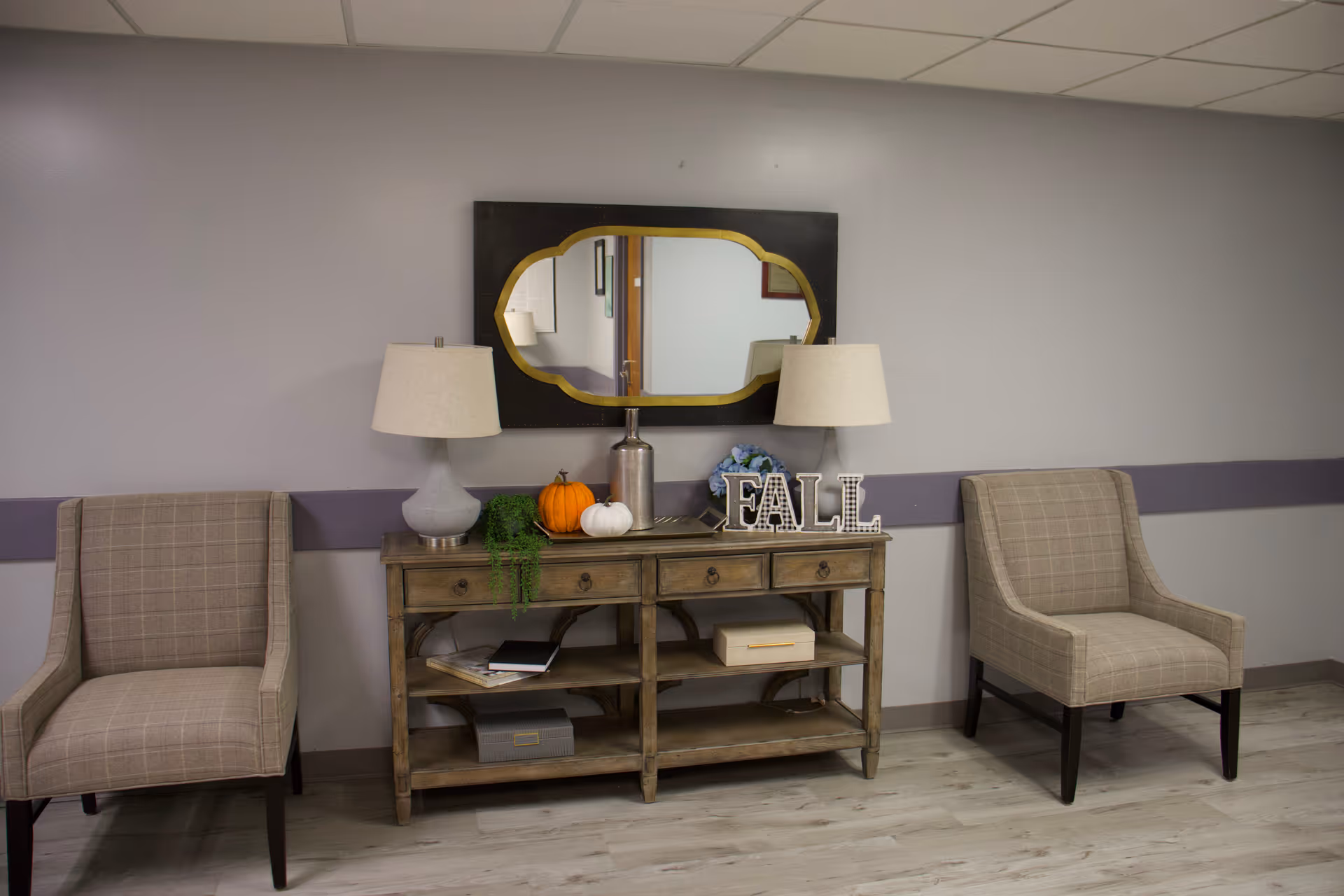 Two beige armchairs flank a wooden console with lamps, pumpkins, and a decorative mirror.