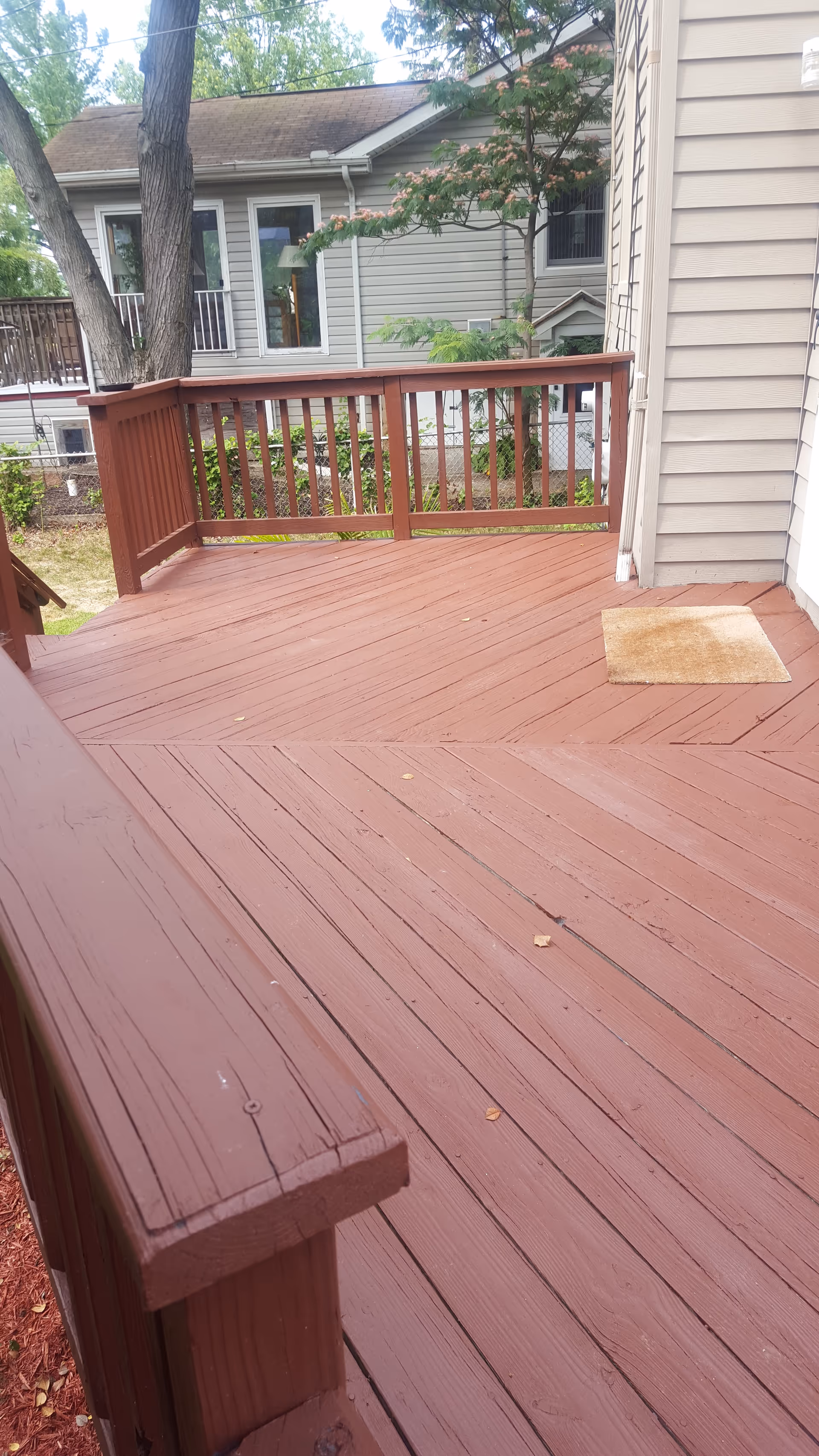 A wooden deck with railings extends from a beige house, trees in the background.