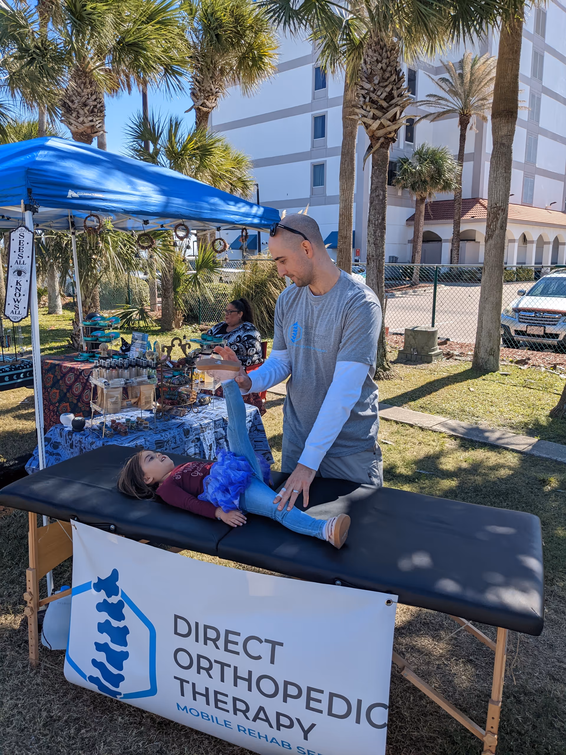 Therapist assists a child on a padded table under a blue canopy outdoors.