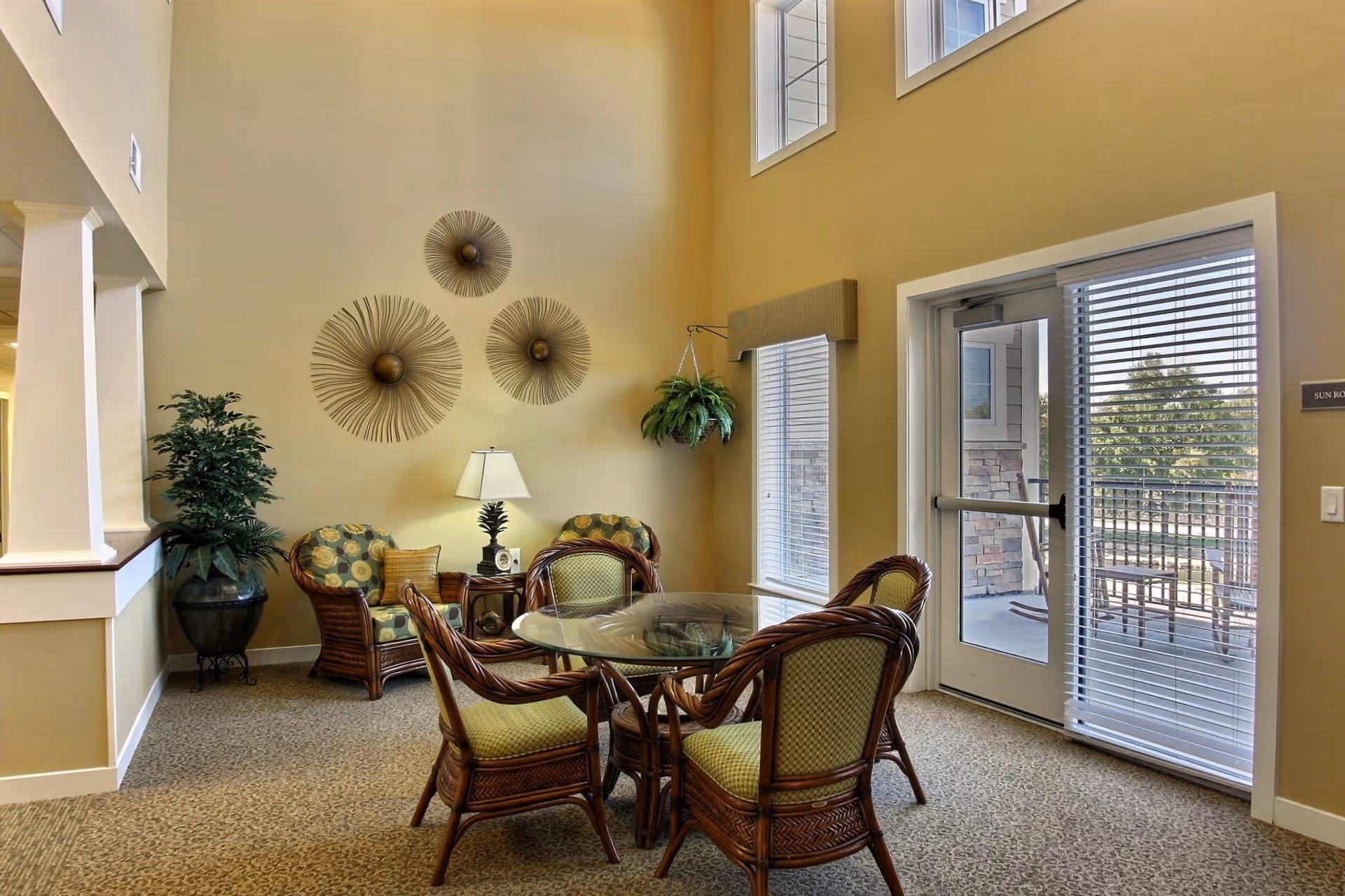 Sunlit senior living common room with wicker chairs around a glass table, decorative wall sculptures and a glass door to a patio.