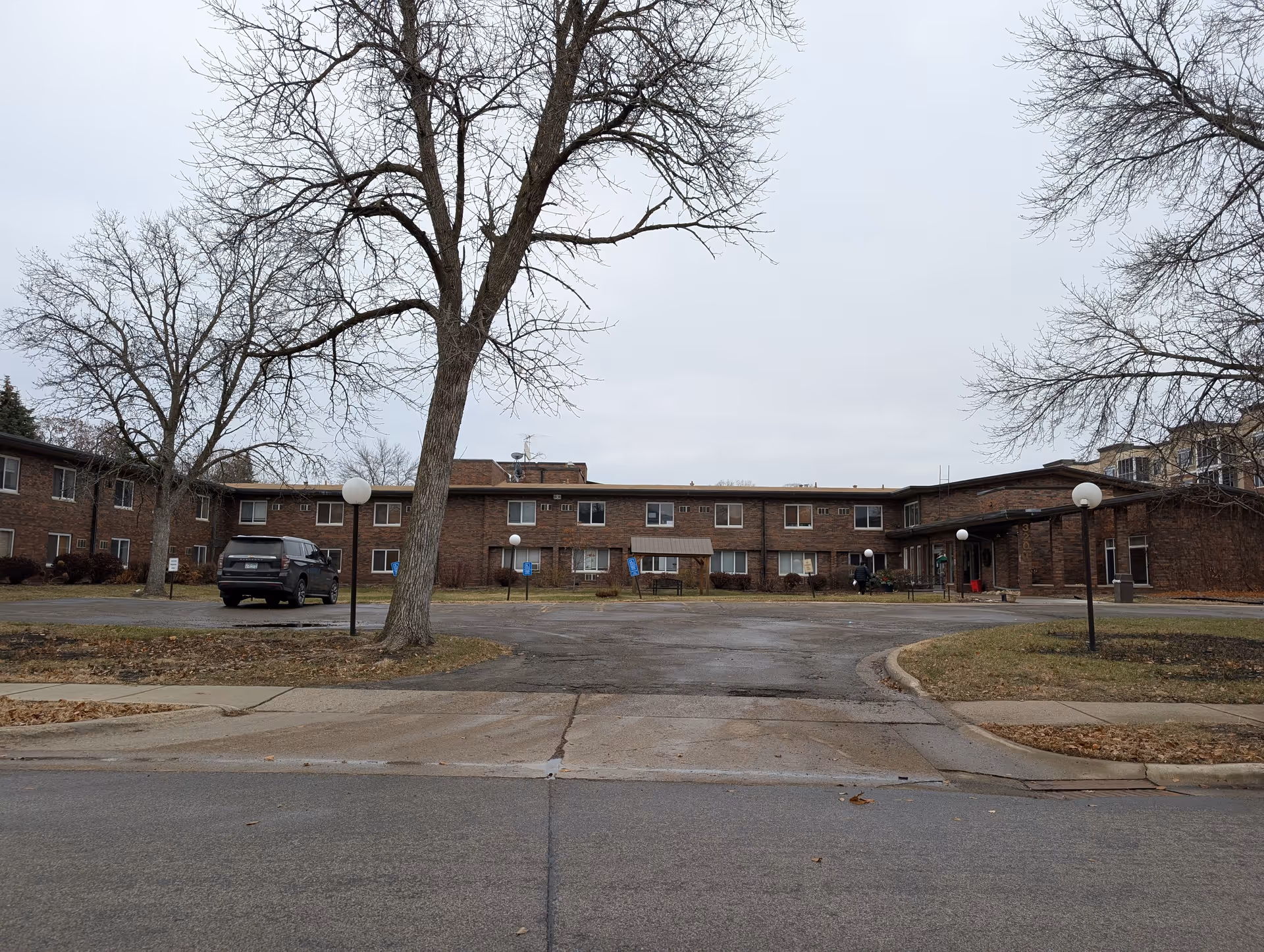 Two-story brick building front with a circular driveway, leafless trees, parked car, and globe lamp posts under an overcast sky.