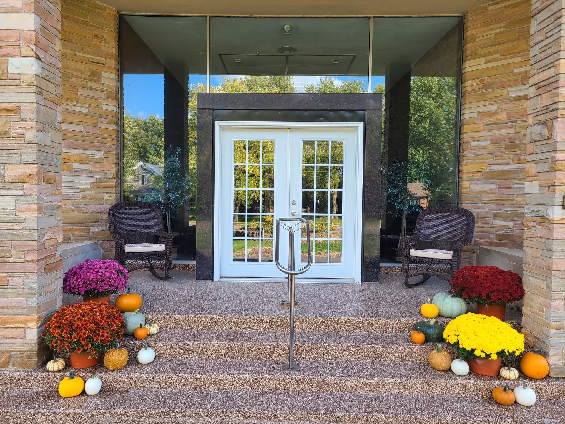 Front entrance with double glass doors, wicker chairs on either side, and potted mums and pumpkins arranged on the steps.