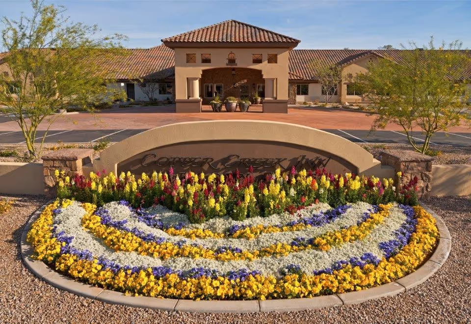 Entrance of Copper Creek Inn Memory Care facility with a curved sign surrounded by colorful flower beds including yellow, purple, white, and red flowers. The building has a tiled roof and a covered entryway with potted plants. Trees and a parking area are visible in the background.