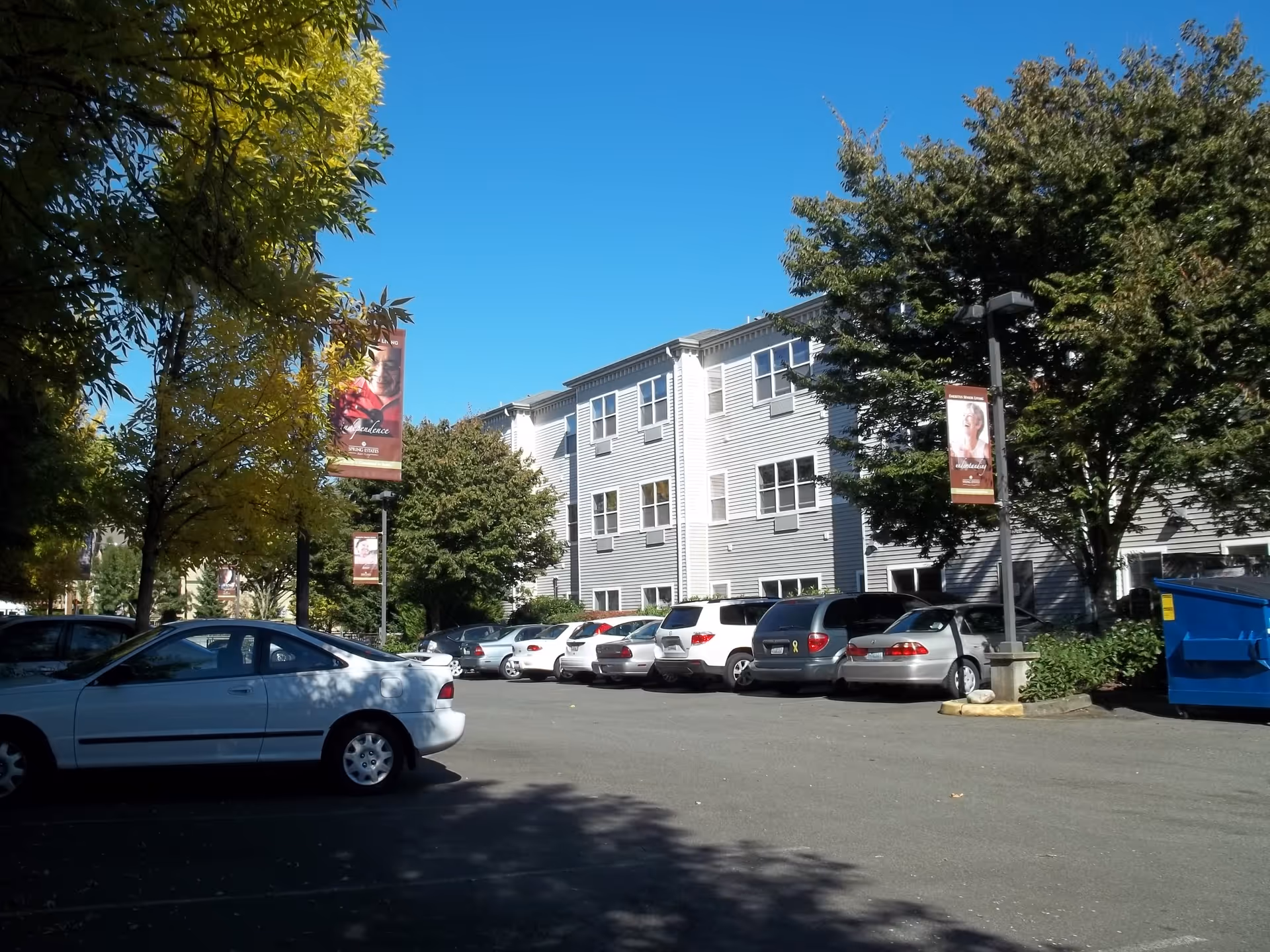 Exterior view of Kenmore Senior Living with parked cars and trees along a sunny day.