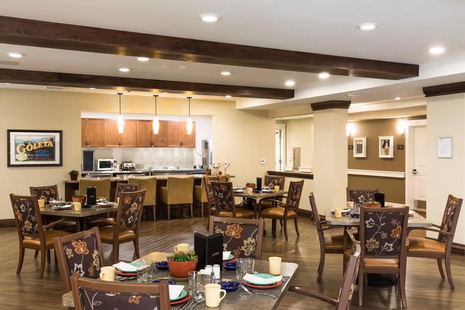 Dining room with several set tables and patterned chairs, a serving counter and kitchenette beneath wooden ceiling beams.