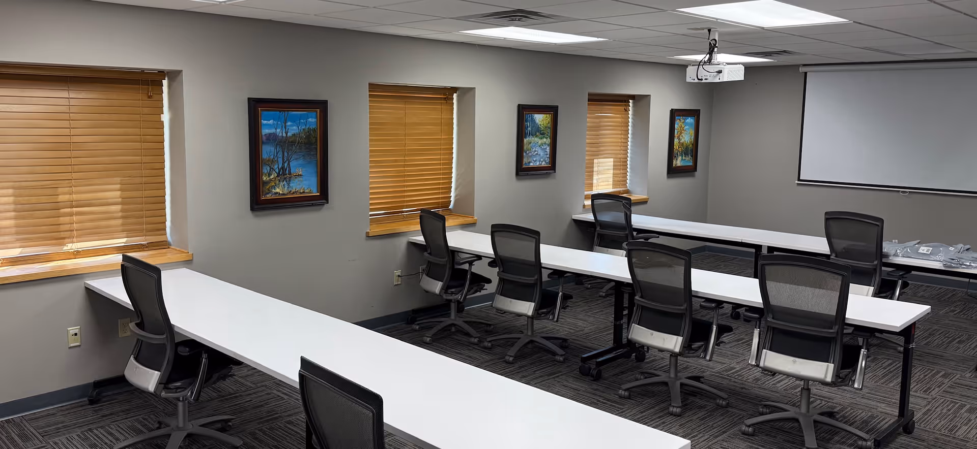 Bright conference room with long white desks, chairs, blinds, windows, and projector.