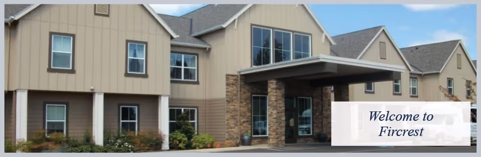 Exterior view of Fircrest Senior Living building showing a two-story structure with beige siding, multiple windows, a covered entrance supported by stone pillars, and some landscaping in front.