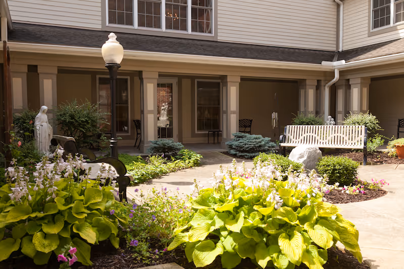 A serene courtyard featuring vibrant flower gardens, a decorative bench, and a lamp post, surrounded by greenery and a building facade.