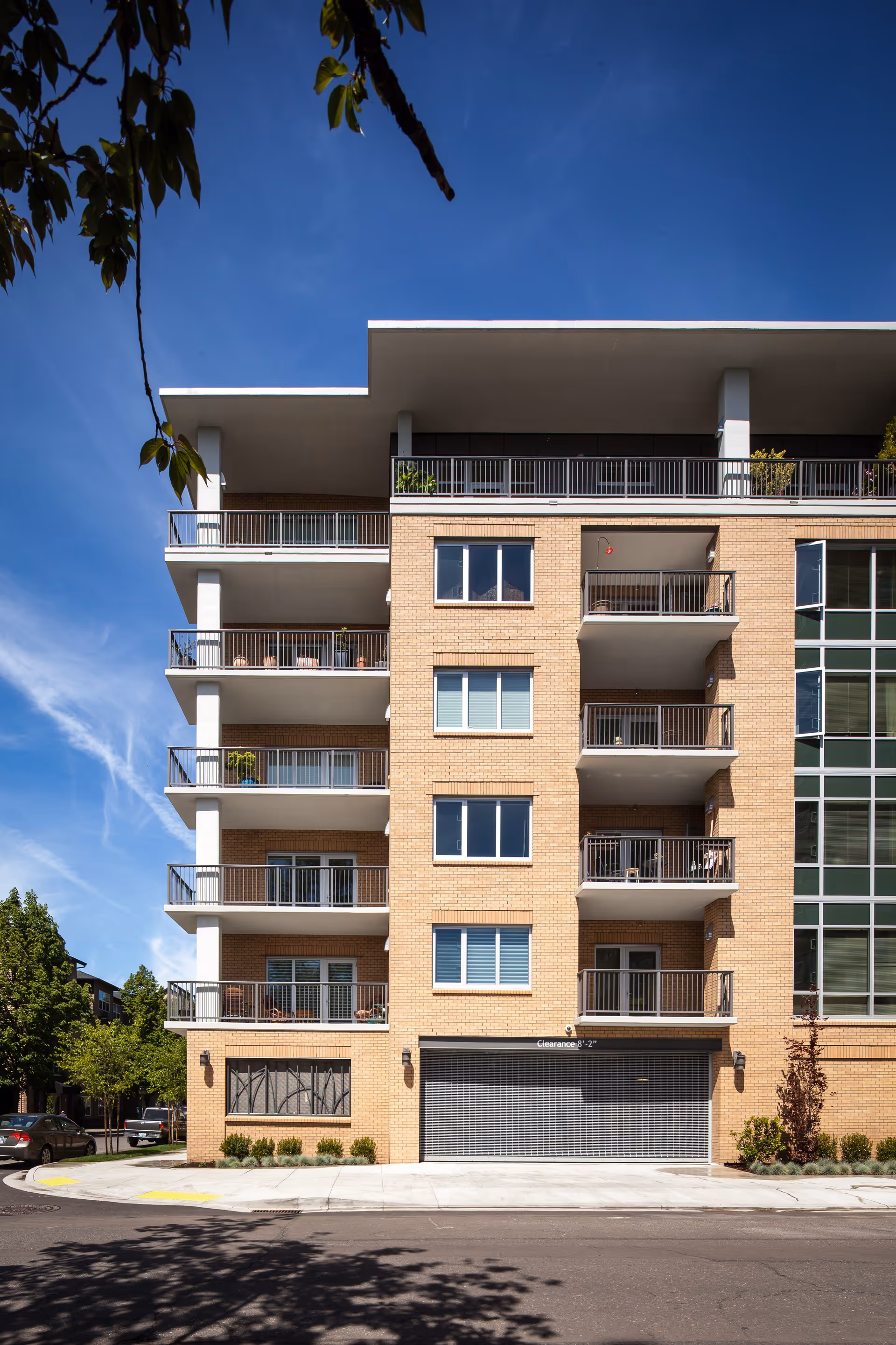 Street-level view of the front facade of a multi-story brick residential building with balconies and a closed garage door under a clear blue sky.