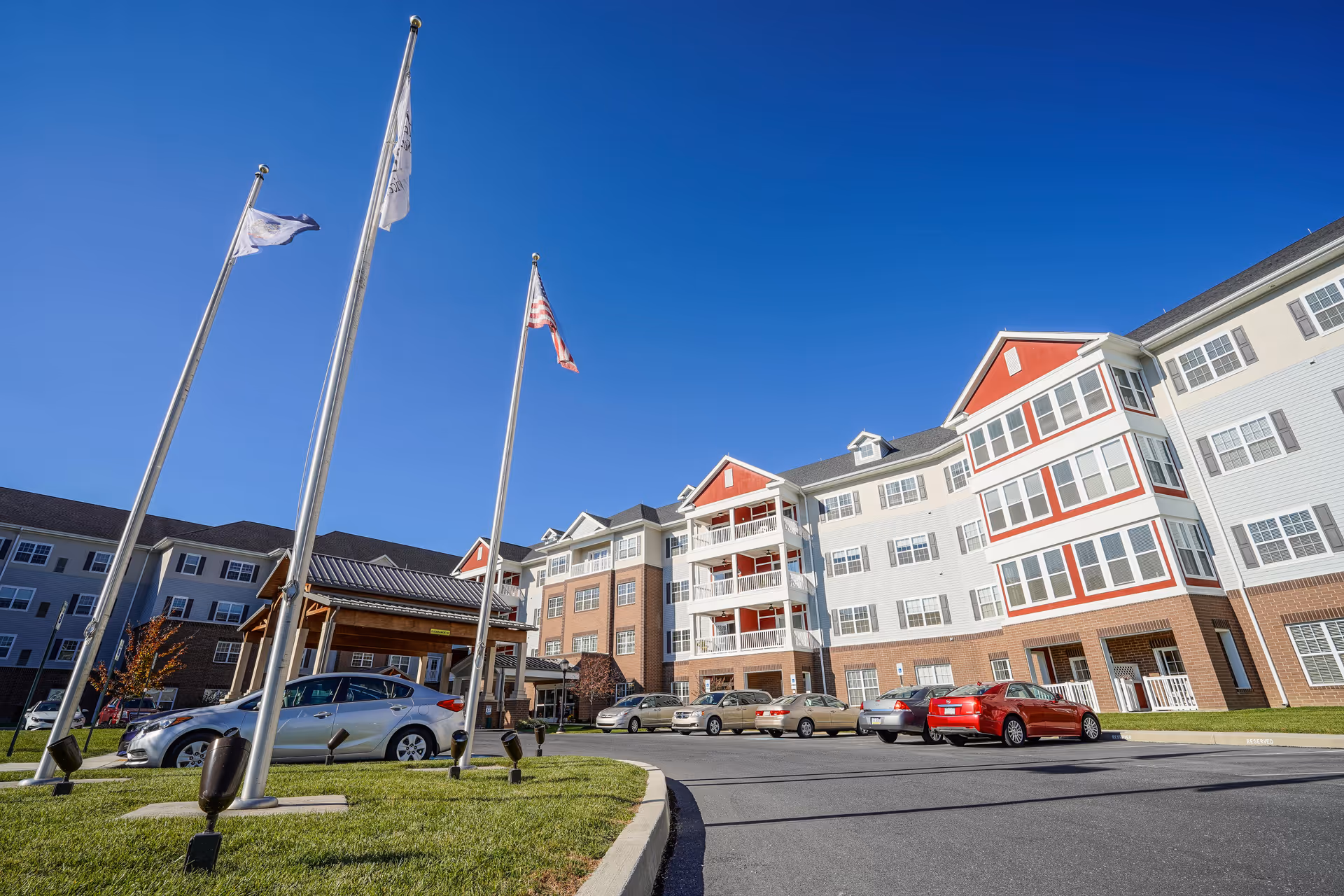 Exterior view of a multi-story senior living building with flagpoles, a covered entrance, and parked cars under a clear blue sky.