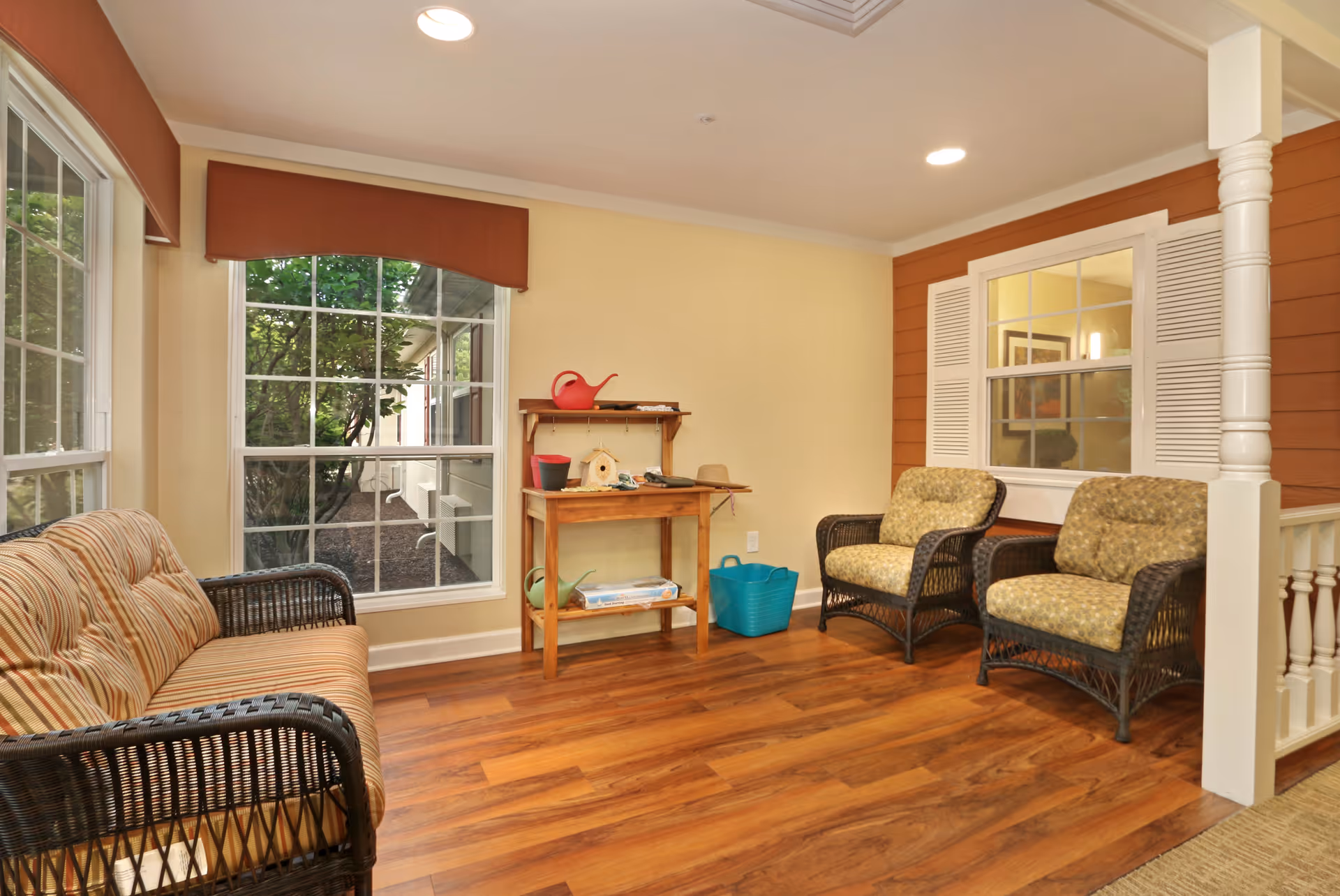 Sunroom-style seating area with a wicker sofa and two chairs, hardwood floor, large windows, and a small table with gardening items.