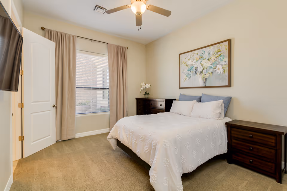 A bedroom with a neatly made bed covered in a white textured bedspread and blue pillows. There are two dark wooden nightstands on either side of the bed, a matching dresser with a white orchid plant, a large floral painting on the wall above the bed, beige curtains covering a window, a ceiling fan with a light, and a door slightly ajar leading to another room.