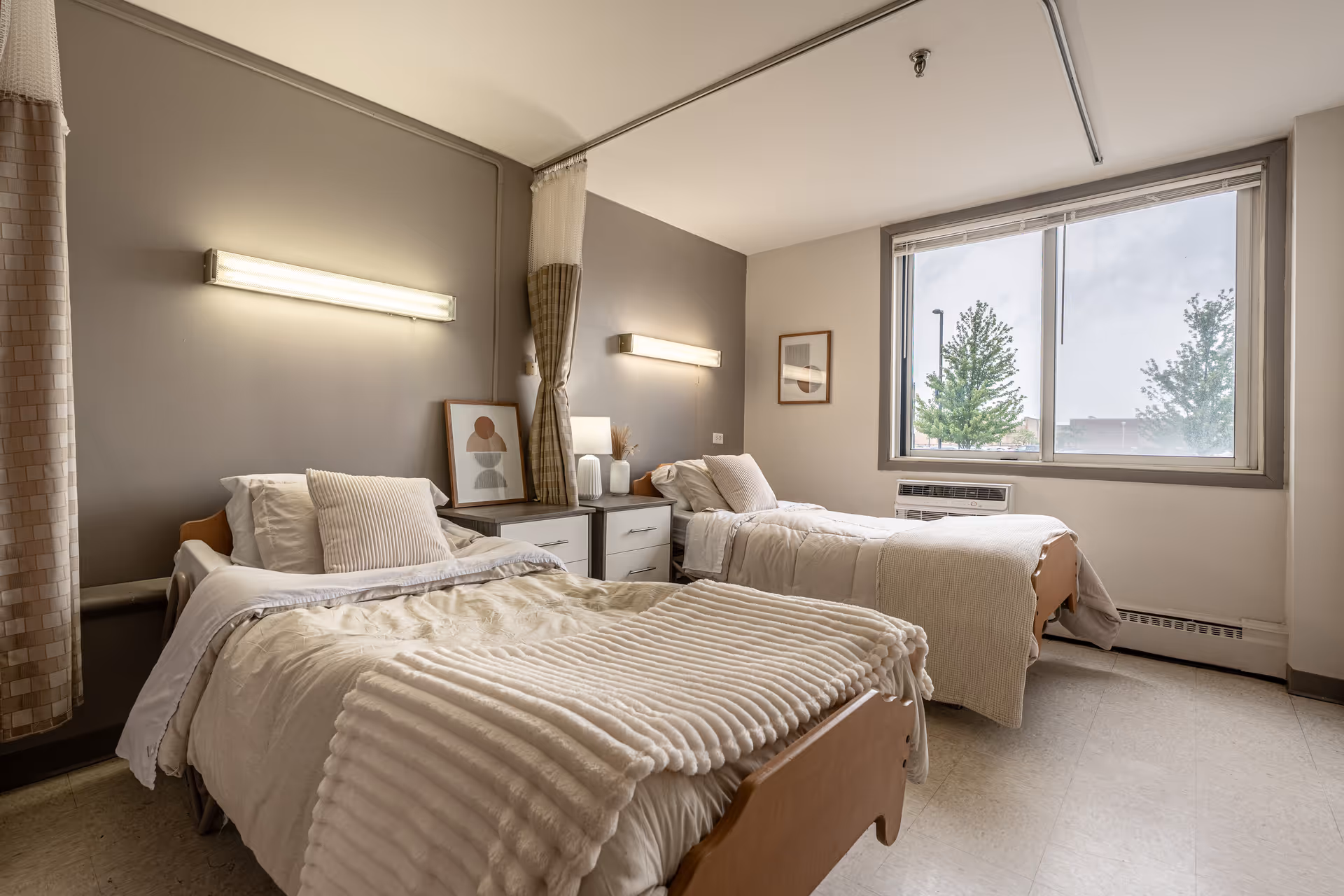 Two twin beds in a neutral, well-lit senior living bedroom with a large window.