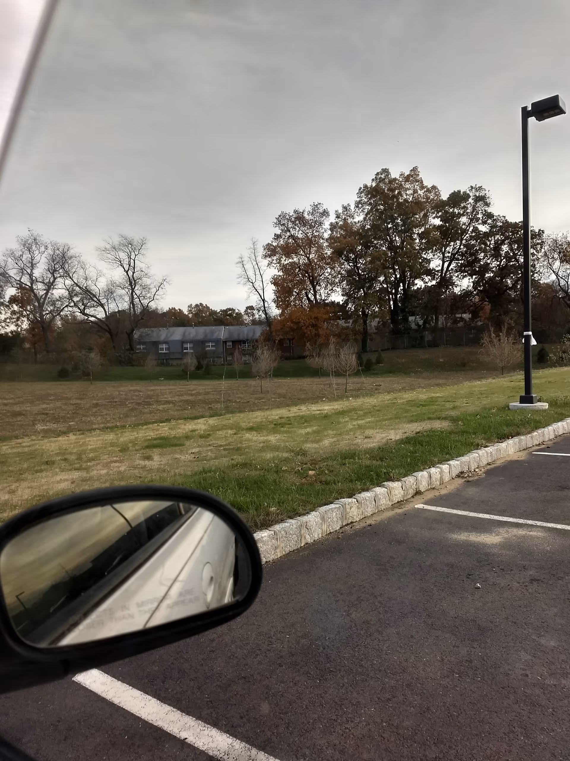 Parking lot with grassy field, trees, and a distant building under a cloudy sky.
