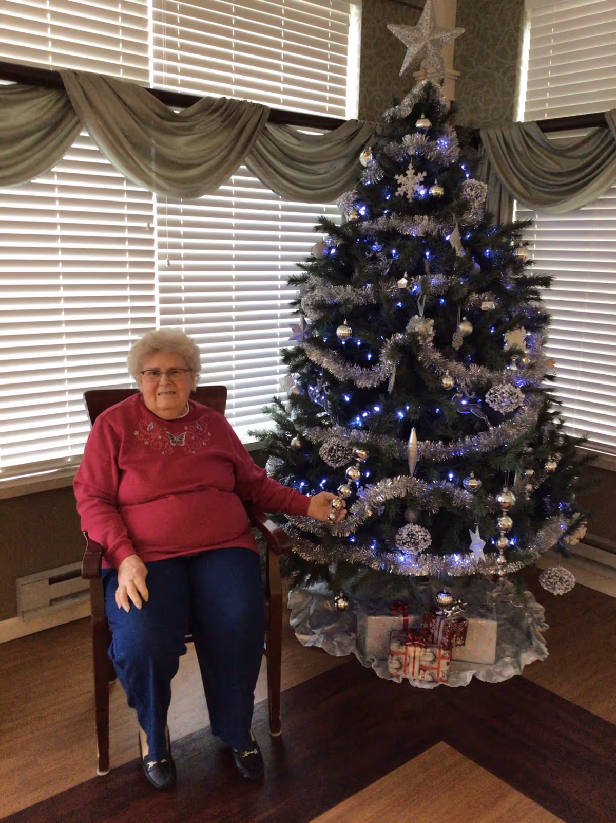 An elderly woman with white hair and glasses sitting on a wooden chair next to a decorated Christmas tree with silver garlands, ornaments, and blue lights. There are wrapped presents under the tree, and the background features large windows with white blinds and green draped valances.