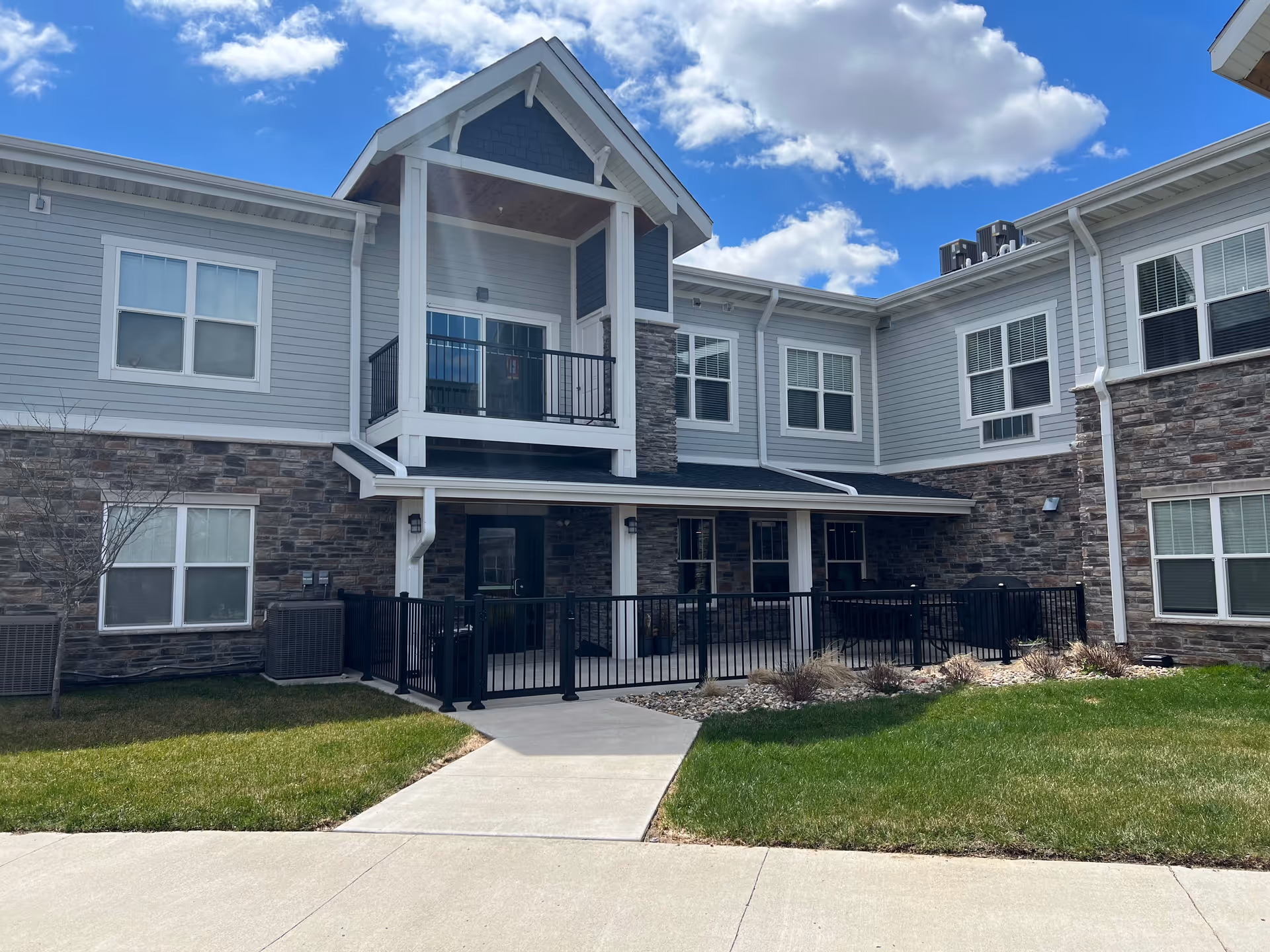 Exterior view of a two-story senior living facility building with gray siding and stone accents. The building features multiple windows, a covered porch area with black railings, and a small balcony above. The sky is blue with scattered clouds, and there is a well-maintained lawn and sidewalk in front.