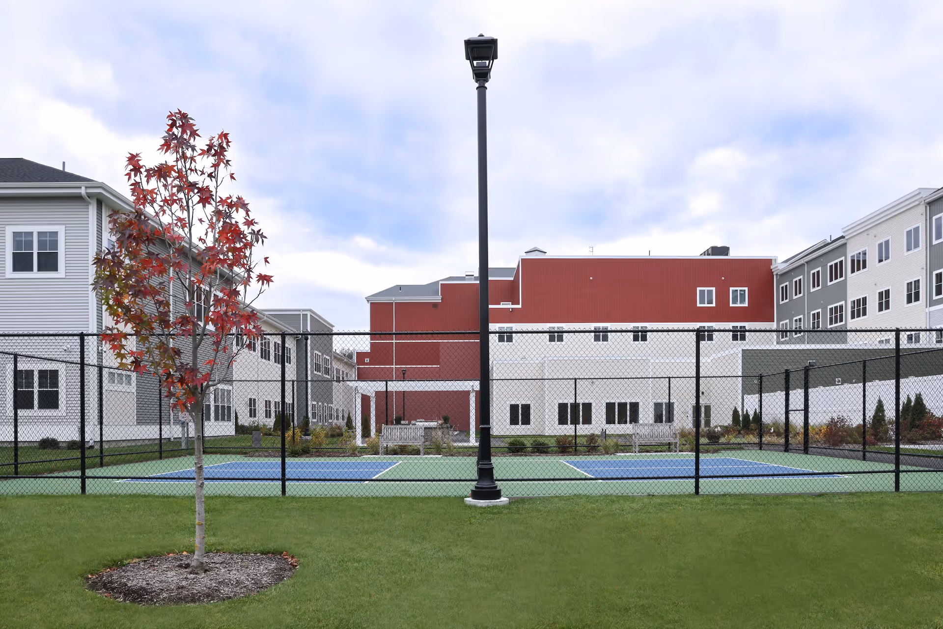 Fenced outdoor tennis courts with a central lamppost, a small red-leafed tree in the foreground, and multi-story retirement buildings behind.