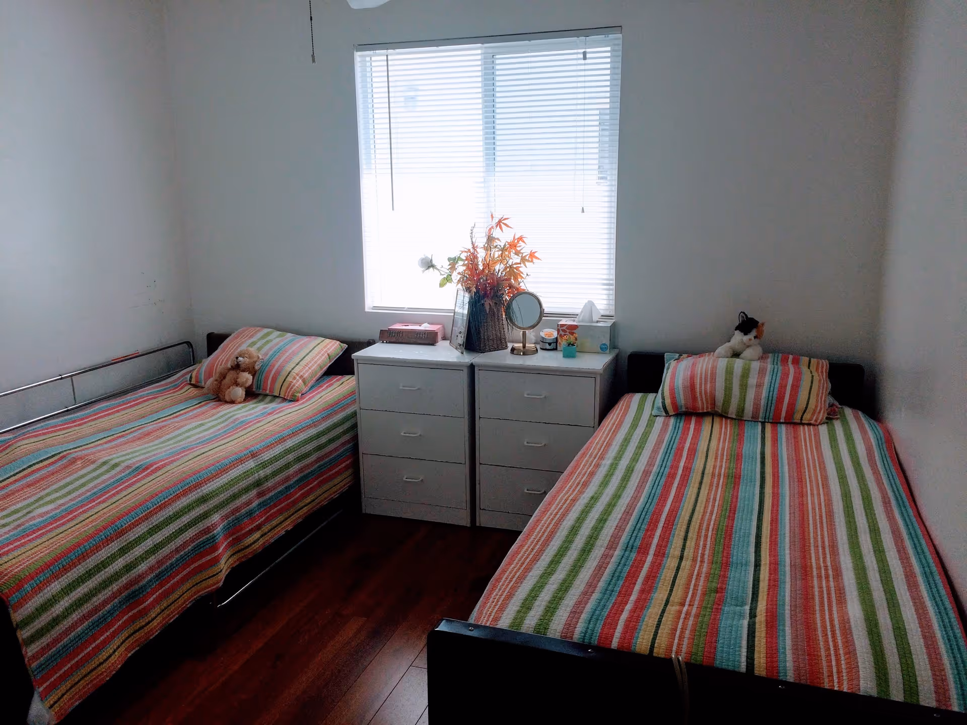 A small bedroom with two single beds placed parallel to each other against opposite walls. Each bed has a colorful striped bedspread and a stuffed animal on the pillow. Between the beds are two white drawer units with a vase of flowers, a small mirror, and other small items on top. A window with closed blinds is centered on the wall above the drawer units. The room has white walls and wooden flooring.
