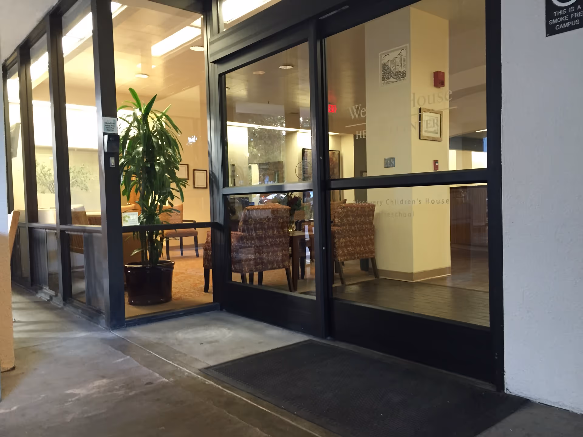 Glass storefront entrance to Webster House lobby with chairs and a tall potted plant inside.