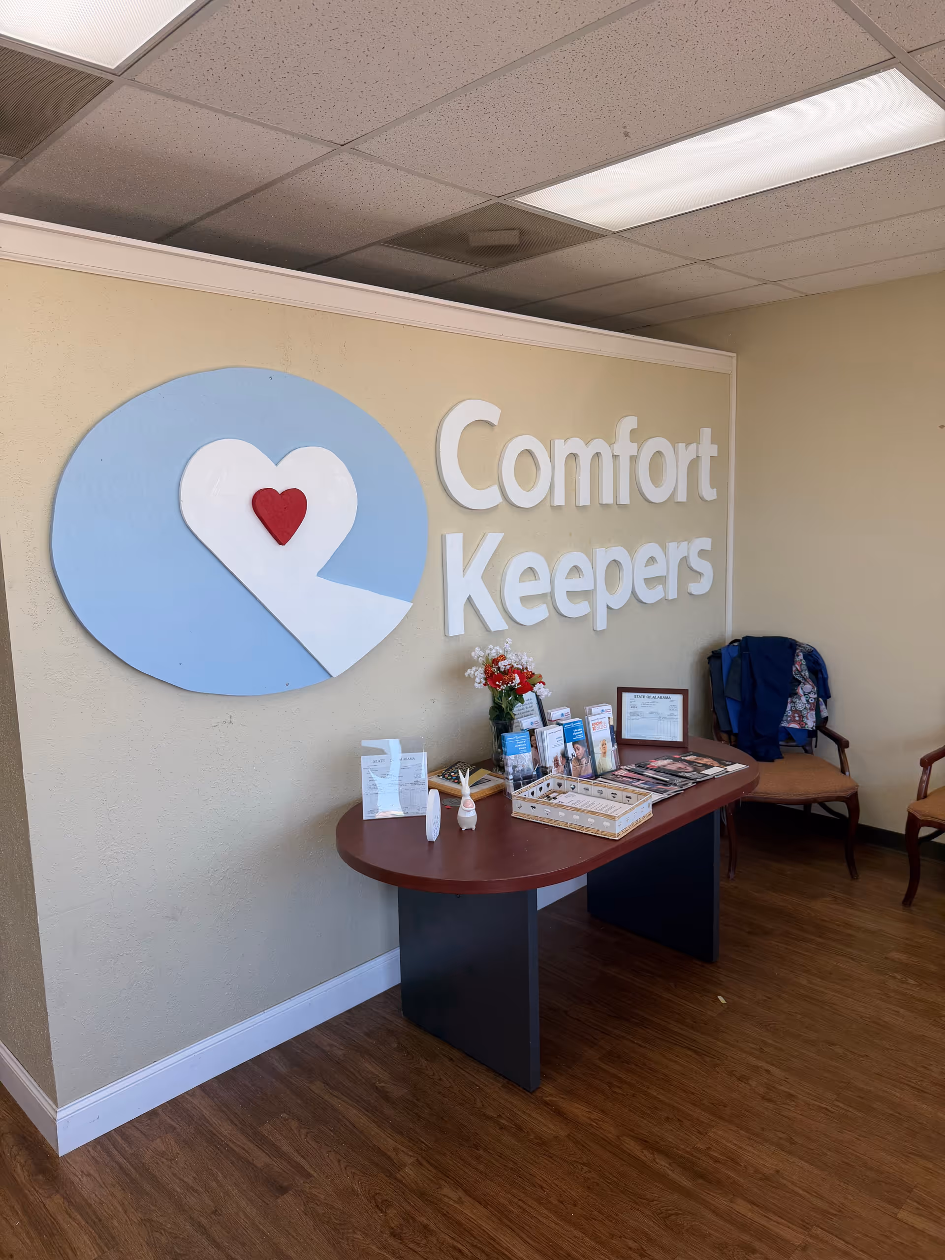 Interior reception area with Comfort Keepers logo, brochures, and flowers on a curved desk.