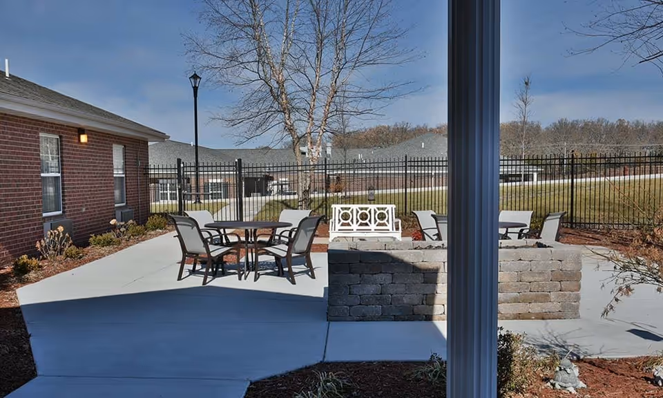 Outdoor patio area at NorthRidge Place with round tables and chairs, a white bench, a brick fire pit, surrounded by a black metal fence and landscaping. The sky is clear and blue with some trees and buildings in the background.