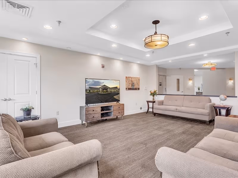 A spacious senior living facility common area with beige sofas arranged around a flat-screen TV on a wooden stand. The room has a neutral color palette with beige walls, carpeted floor, recessed lighting, and a decorative ceiling light fixture. There are side tables with flowers and a globe, and a double door on the left side of the room.