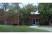 Brick-front senior living building entrance with glass doors, trees, and a grassy lawn