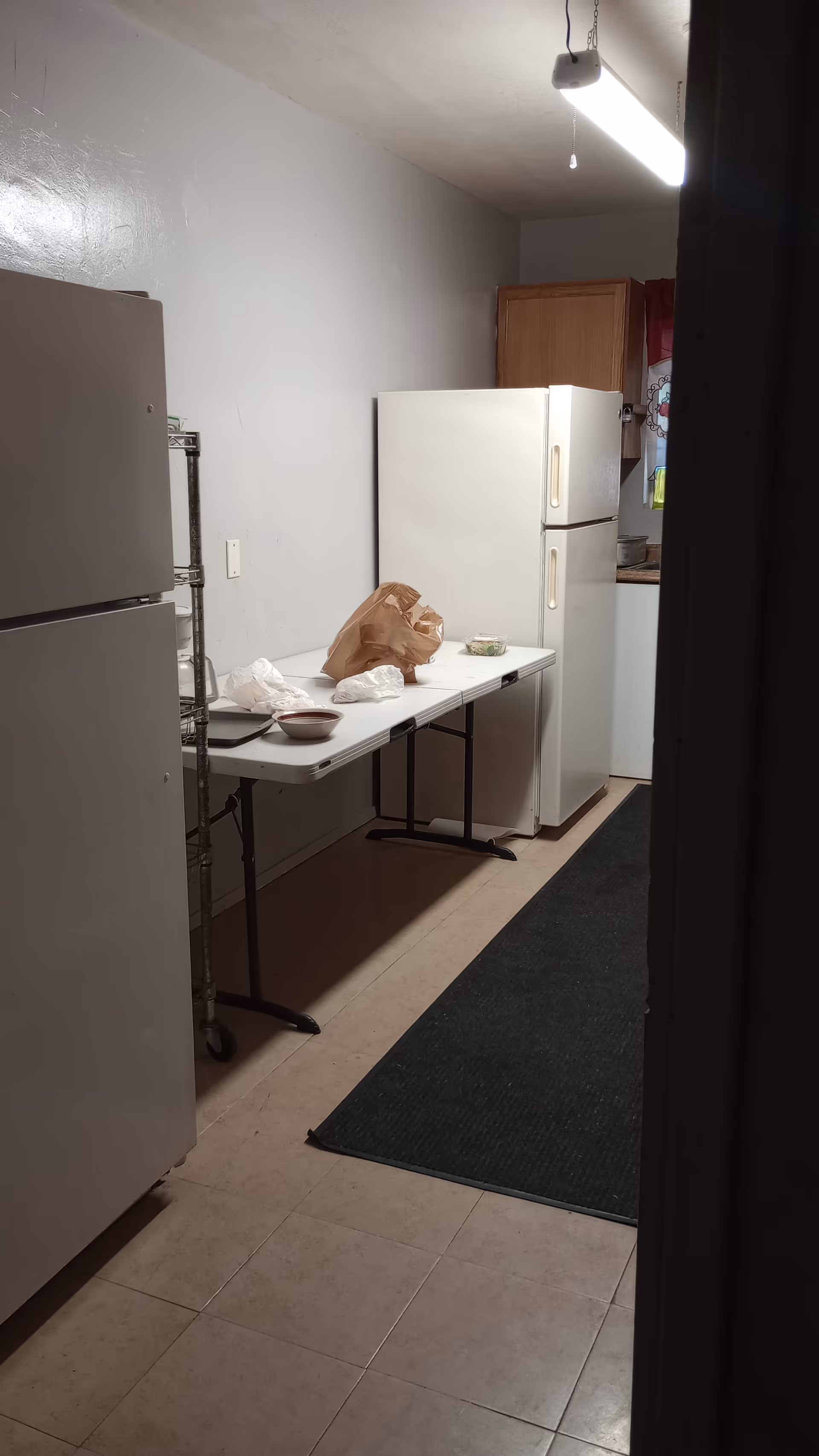 Narrow kitchen in a senior facility with a fridge, table, and bags.