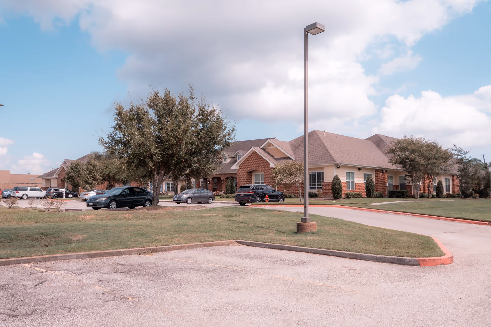Single-story brick senior living building with a parking lot, trees, and a lamp post under a partly cloudy sky.