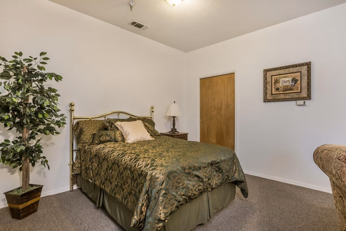 A bedroom with a bed covered in green and gold patterned bedding, a wooden nightstand with a lamp, a potted artificial plant, a framed picture on the wall, and a closed wooden door.