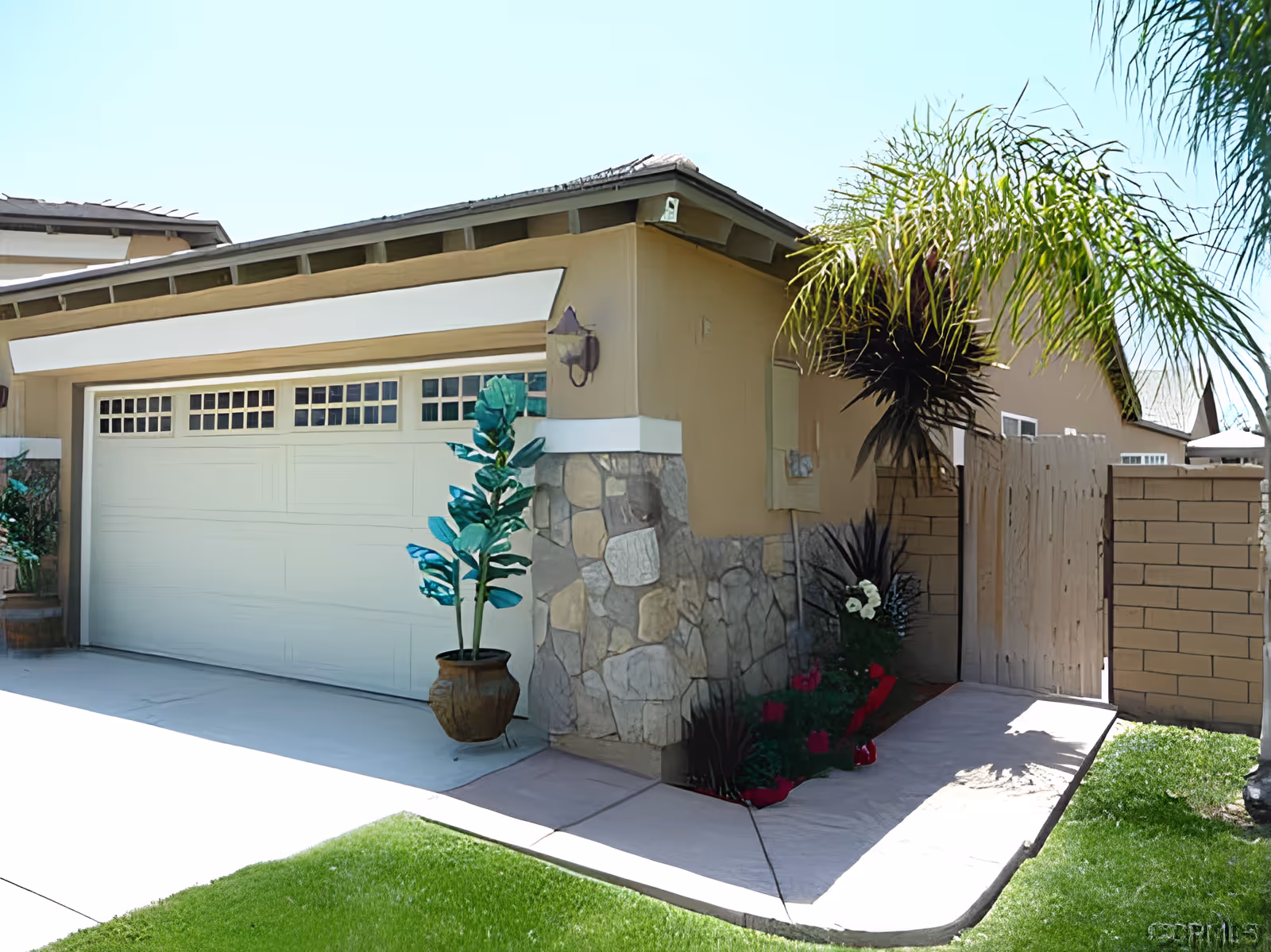 Front exterior of a house showing a closed garage door, potted plants, stone-clad wall, palm tree, and a side gate.