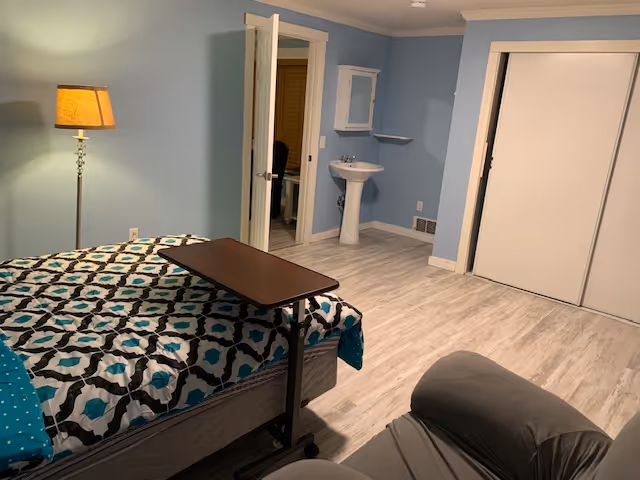 Bedroom in a senior facility with a patterned bed, recliner, lamp, and small sink.
