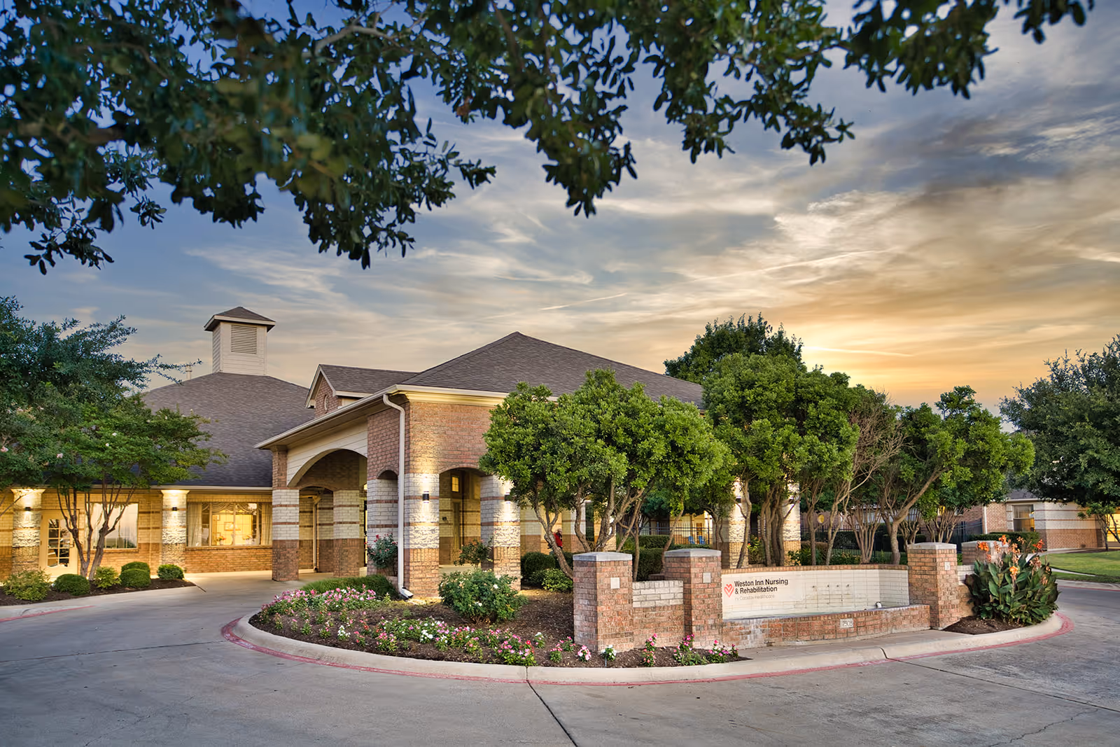 Exterior view of Weston Inn Nursing and Rehabilitation facility at sunset, showing a brick building with a covered entrance, surrounded by trees and landscaped flower beds.