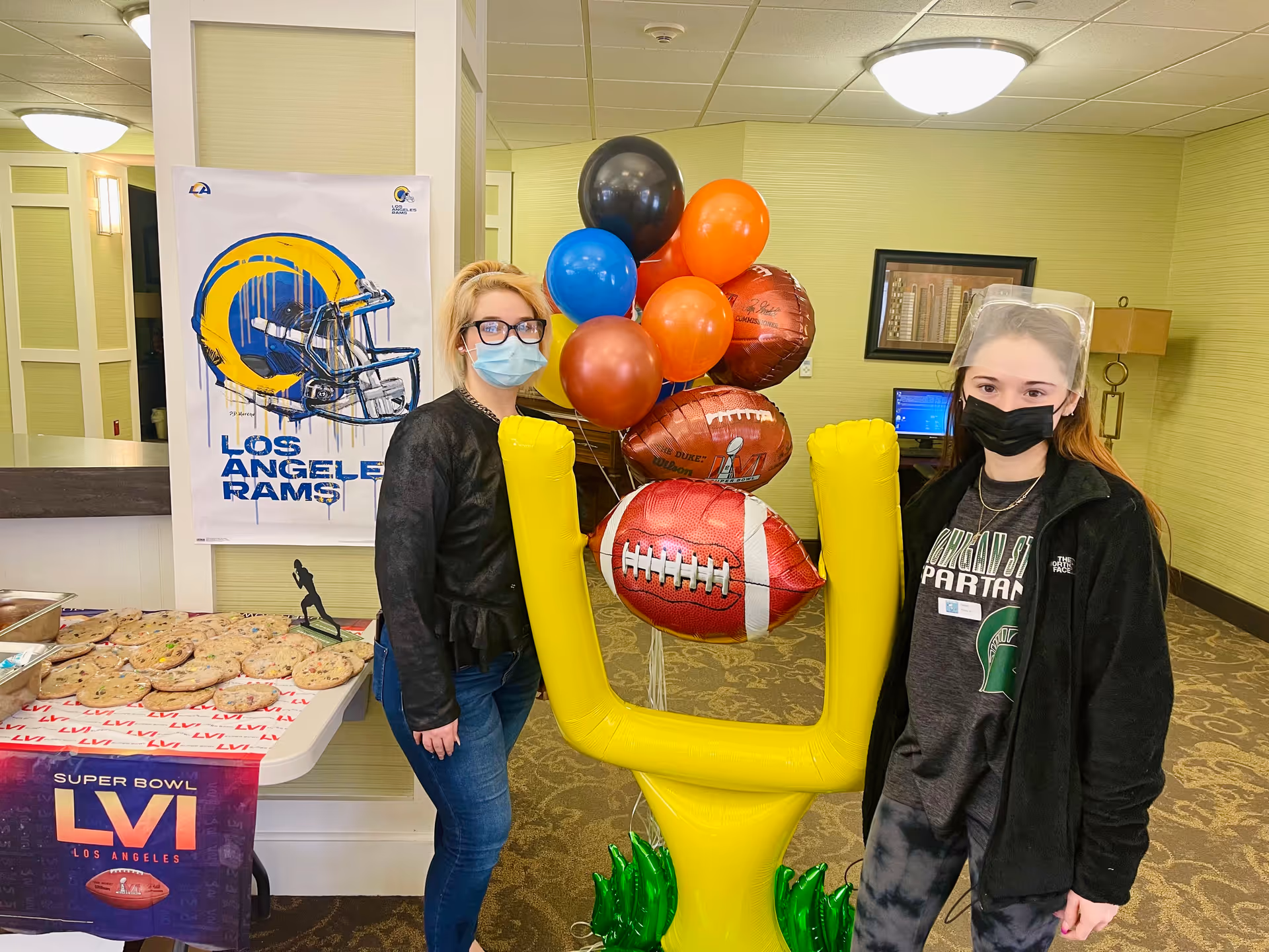 Two women wearing face masks and standing indoors next to a yellow inflatable football goalpost decoration with balloons shaped like footballs. A table with cookies and a Super Bowl LVI banner is visible on the left, along with a Los Angeles Rams poster on the wall behind them.